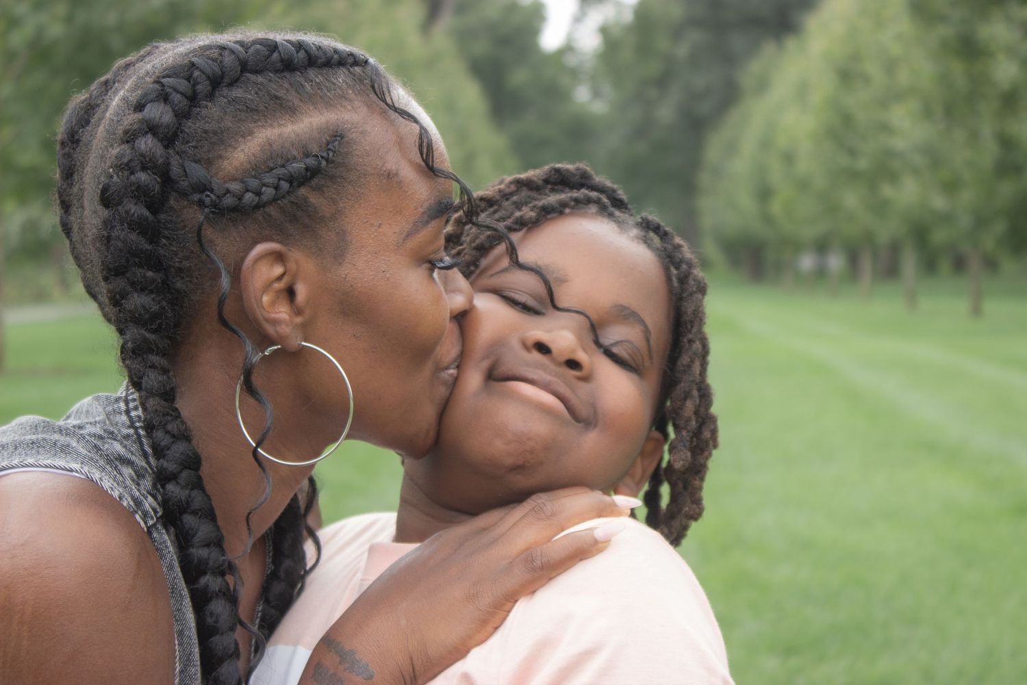 Mother kissing her smiling daughter on the cheek during a tender outdoor family photography session in a green park.