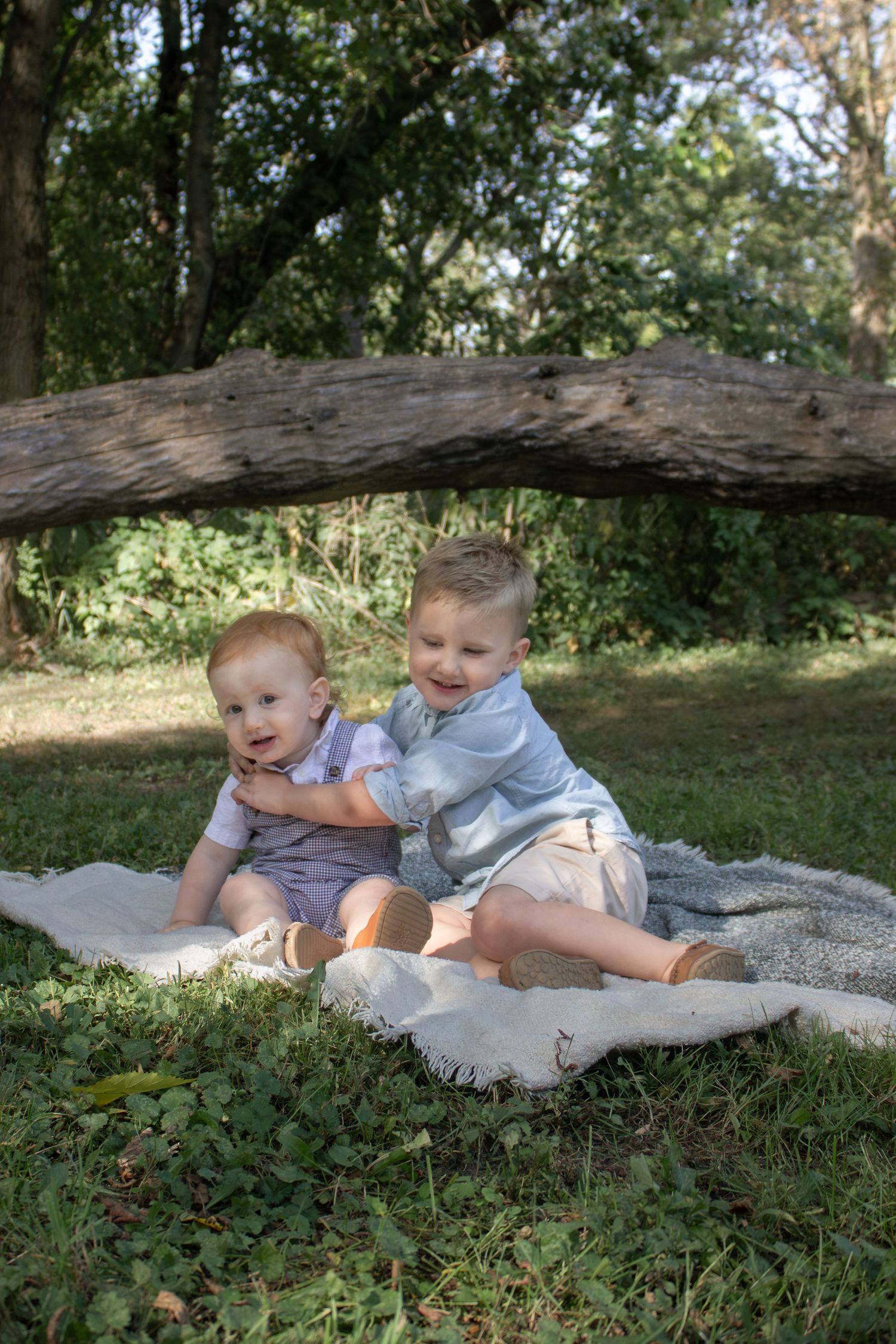 Two young siblings sitting together on a blanket beneath a fallen tree during a natural outdoor children's photography session.