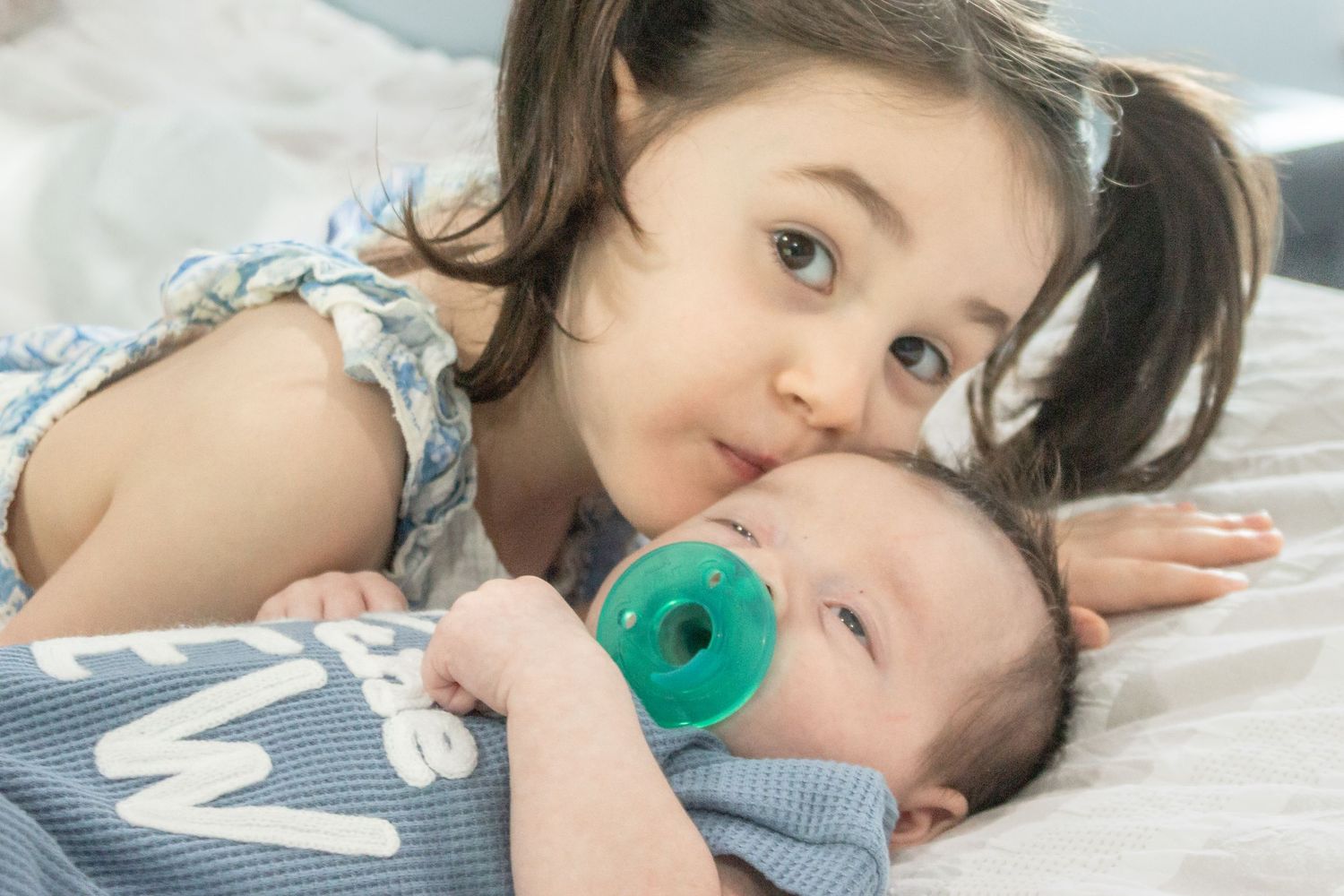Big sister kissing her baby brother on the forehead as he rests with a green pacifier during a natural in-home newborn photo session.