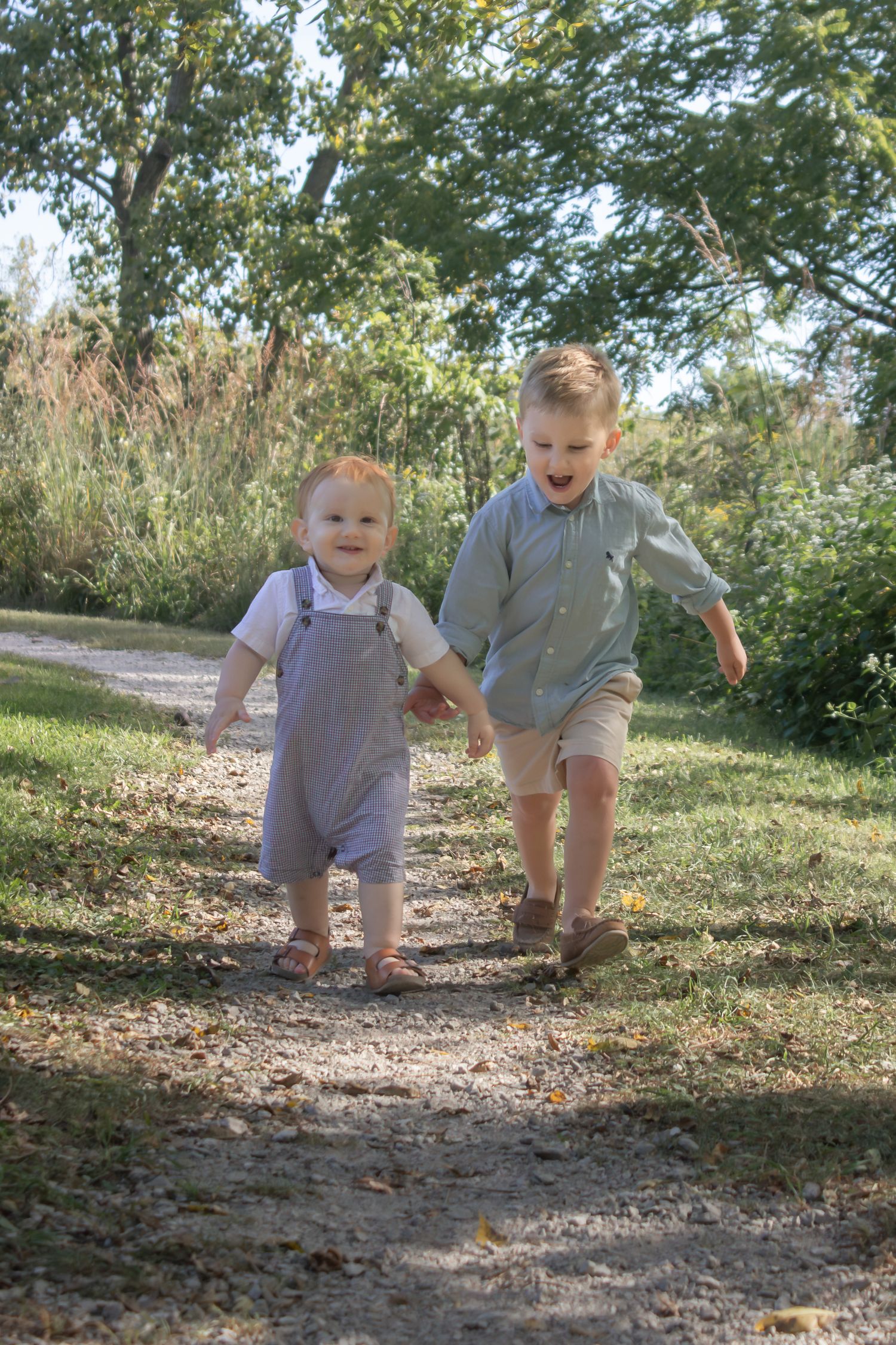 Two young siblings holding hands while walking along a sunlit wooded trail during an outdoor children's photography session.
