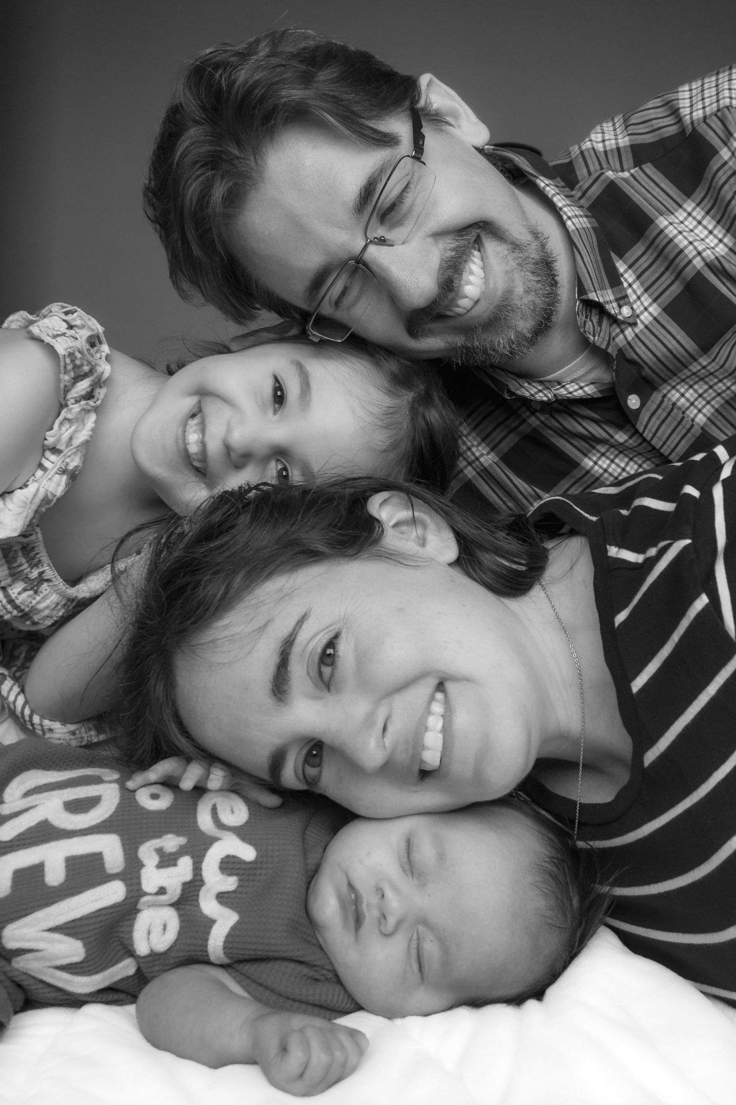 Black-and-white portrait of smiling parents and their young daughter cuddling a newborn baby during an in-home family photo session.