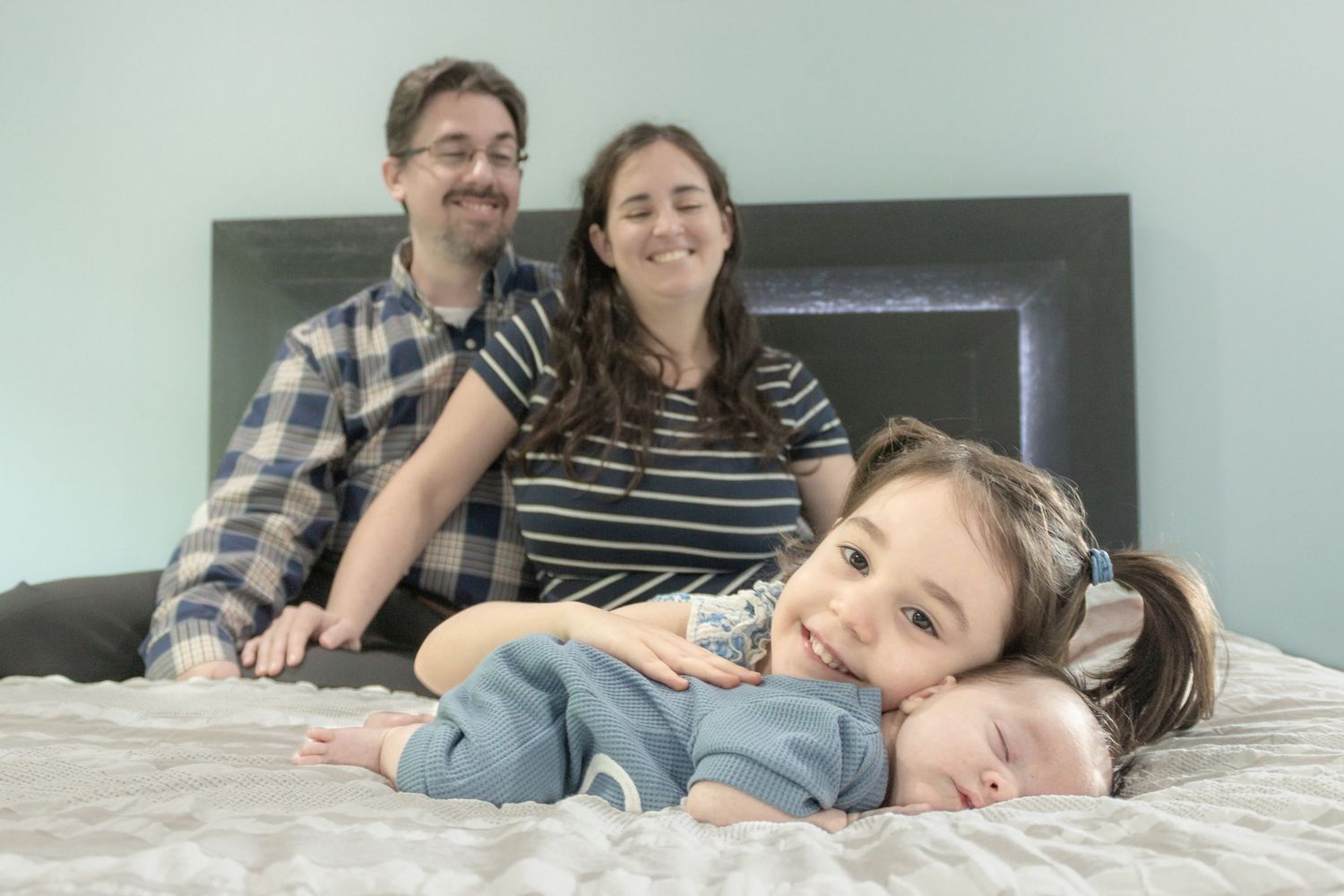 Parents smiling with their young daughter and newborn baby on a bed during a relaxed in-home family photography session.