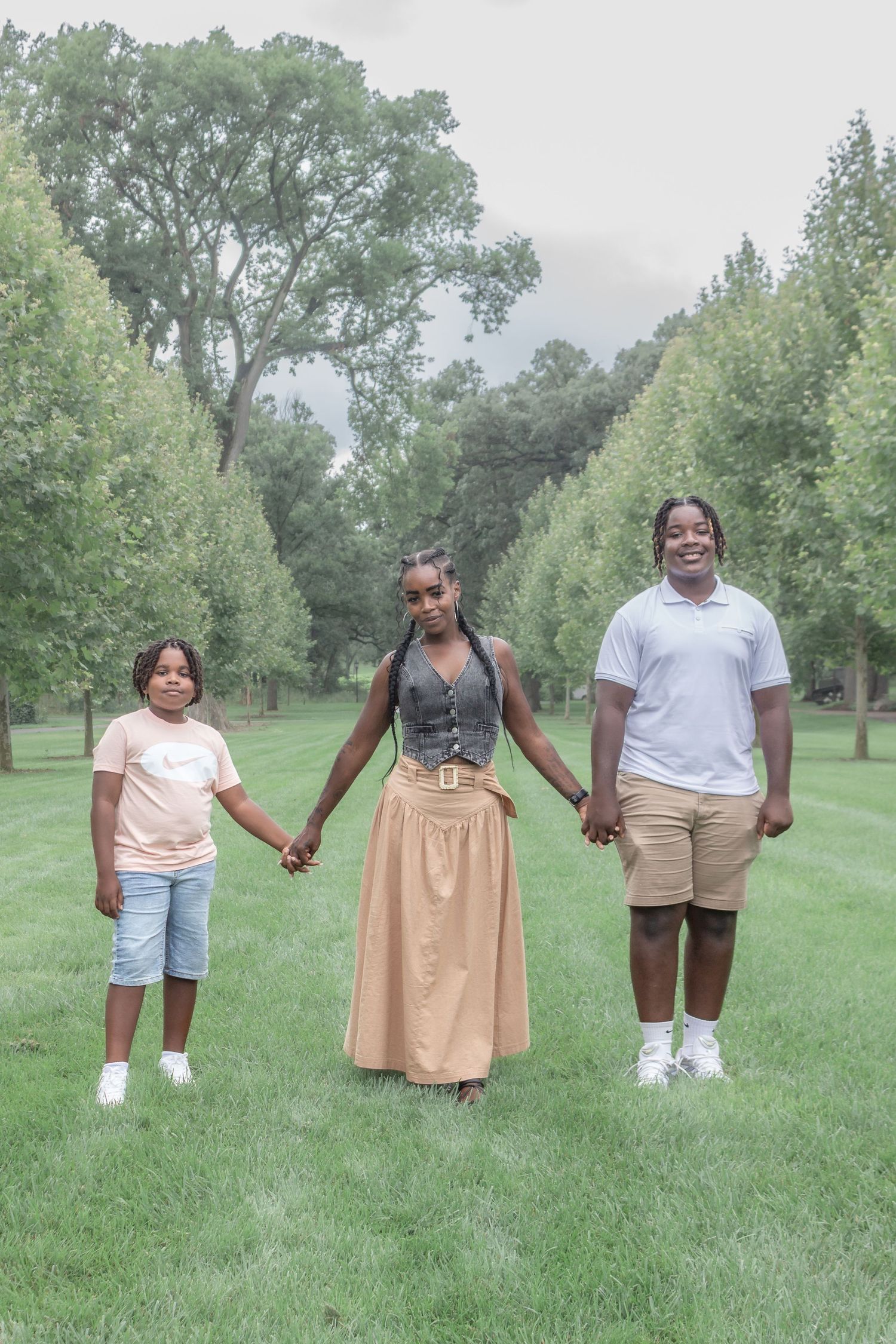 Mother in a tan dress holding hands with her two children on a grassy park pathway during an outdoor family portrait session.