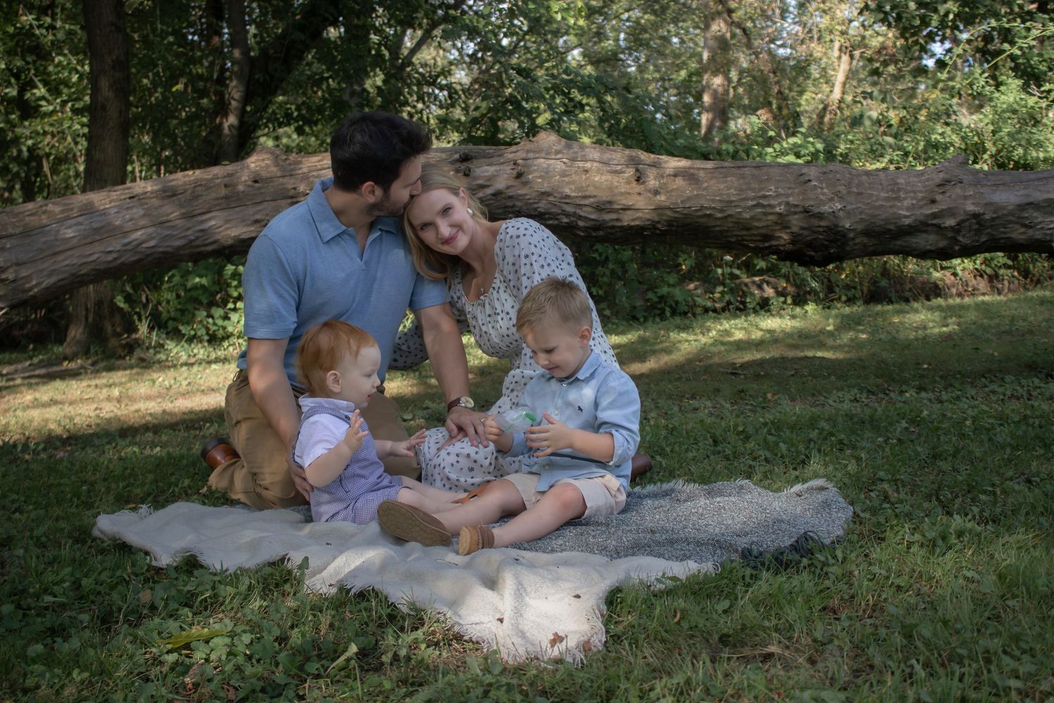 Parents and two young children sitting together on a blanket beneath a fallen tree during a natural outdoor family photography session.