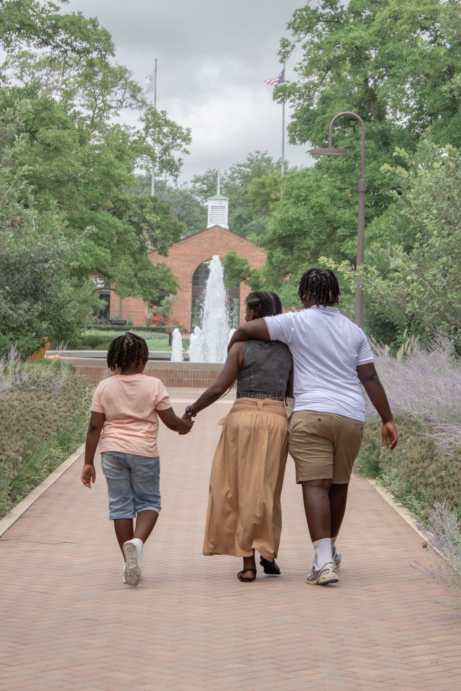 A family of three walks hand-in-hand down a brick pathway toward a fountain in a lush green park.
