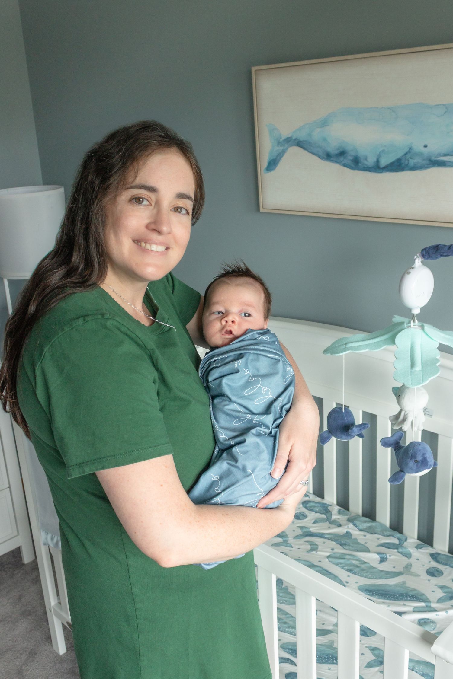 Smiling mother in a green shirt holding her baby beside a white nursery crib during a natural in-home newborn photography session.
