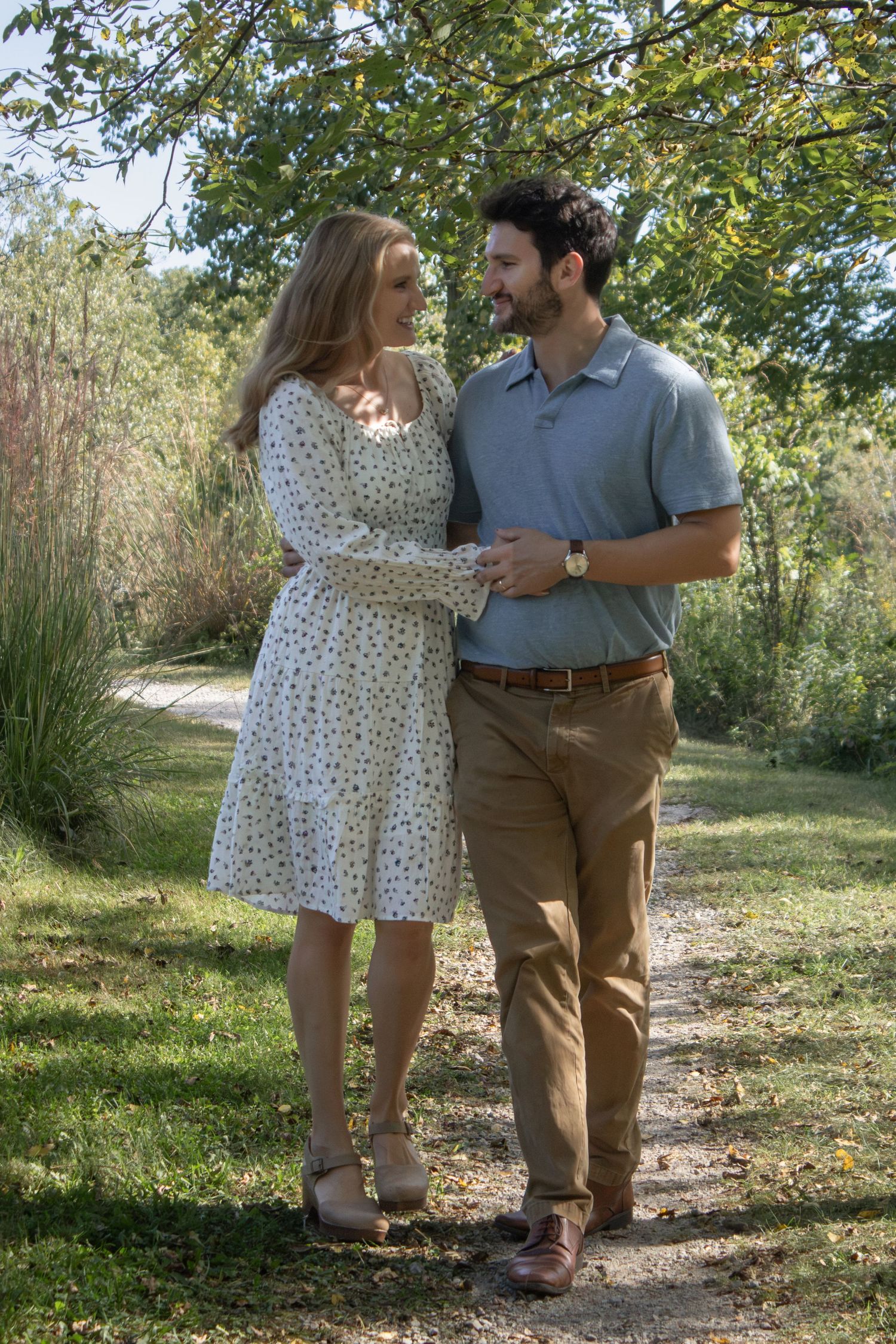 Smiling couple embracing on a sunlit wooded trail during a natural outdoor couples photography session in the Chicago suburbs.