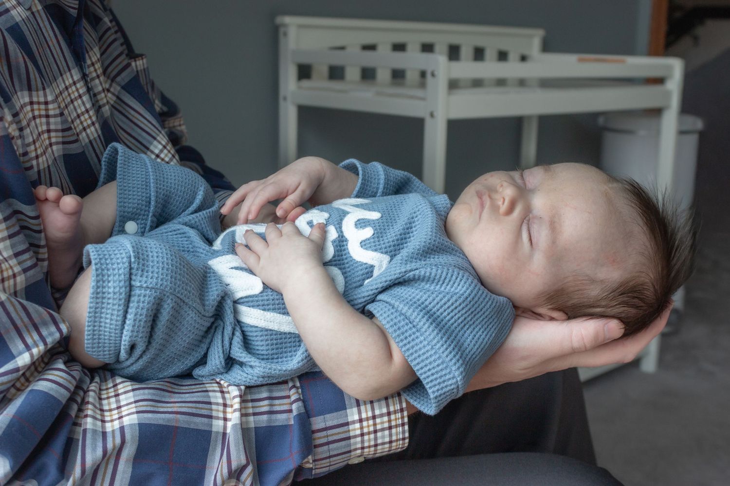Sleeping newborn baby in a blue outfit cradled in a parent's arms during a cozy in-home newborn photography session.