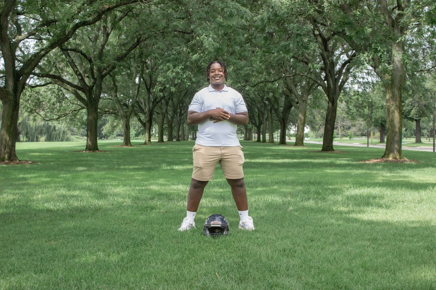 A smiling man stands confidently on a green park lawn with a football at his feet.