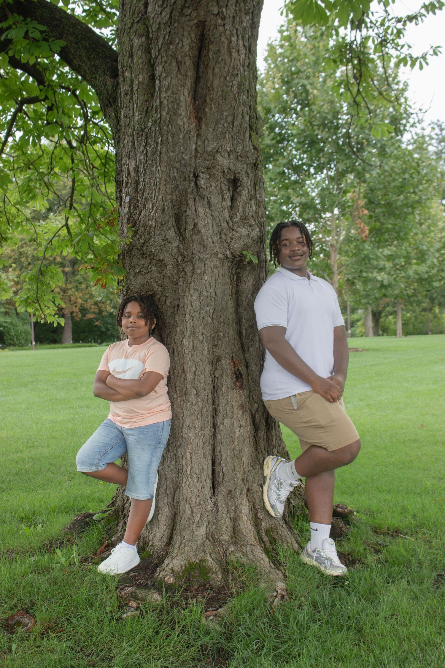 Two siblings posing on opposite sides of a large tree in a green park during a natural outdoor family photography session.