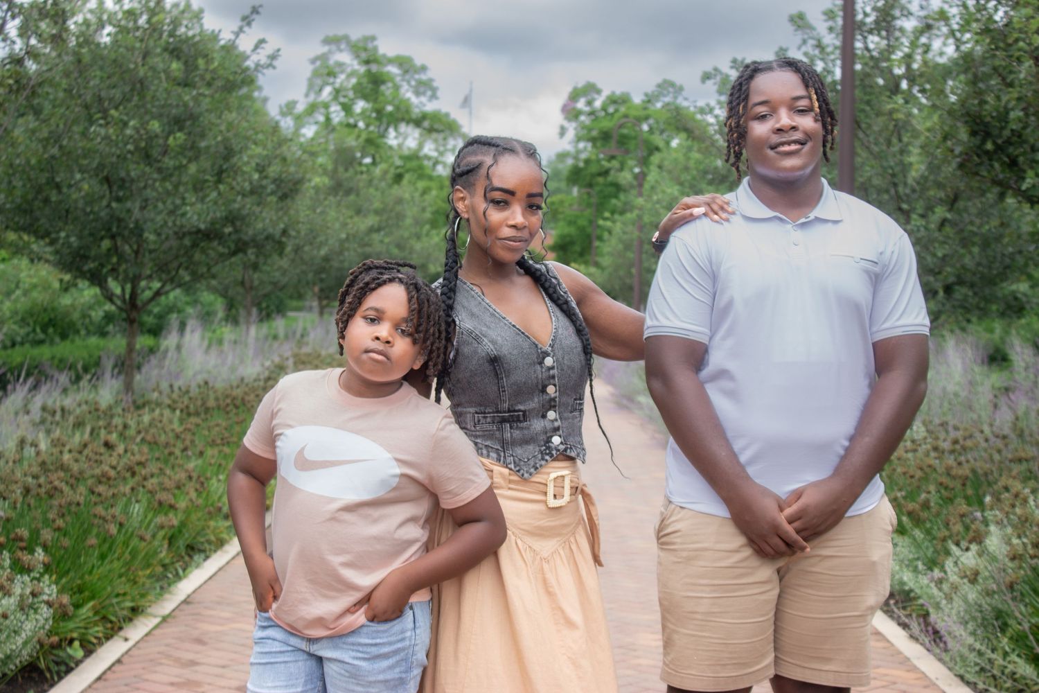 Mother in a tan dress posing with her two children on a tree-lined park pathway during an outdoor family photography session.