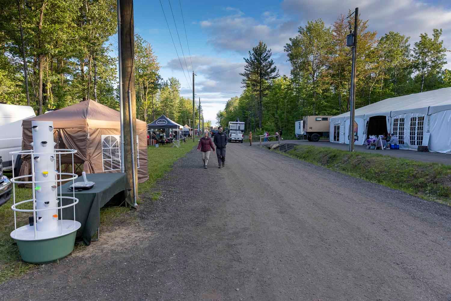 People walking along a dirt road lined with vendor tents and tall trees at an outdoor market fair.
