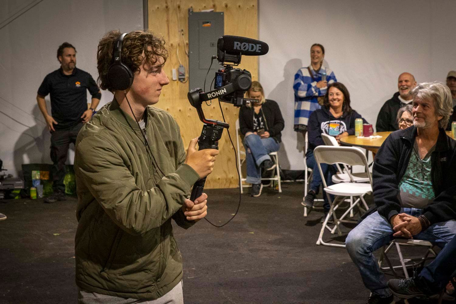Videographer with RODE mic on gimbal stabilizer filming indoor event with audience seated in background.