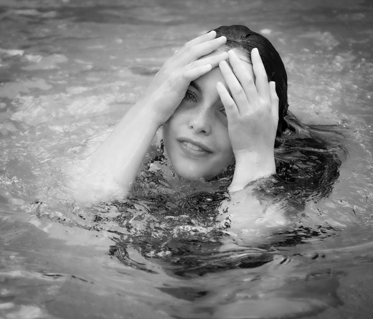 Black and white photo of a smiling woman in a pool, hands raised near her face, surrounded by splashing water.