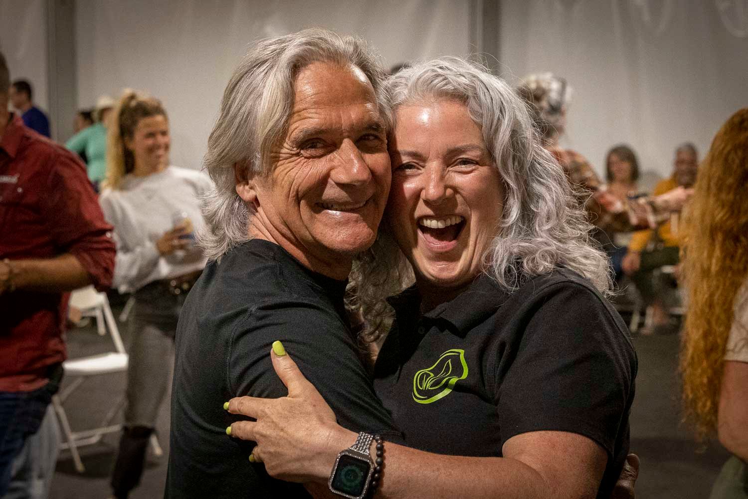 Two people with gray hair share a warm hug and smile at an indoor event, wearing matching black shirts.