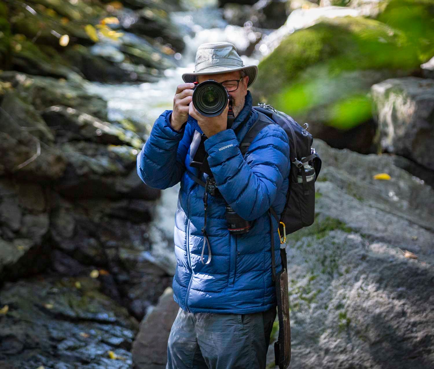 Photographer in blue jacket and hat capturing photos with DSLR camera near rocky stream in forest.