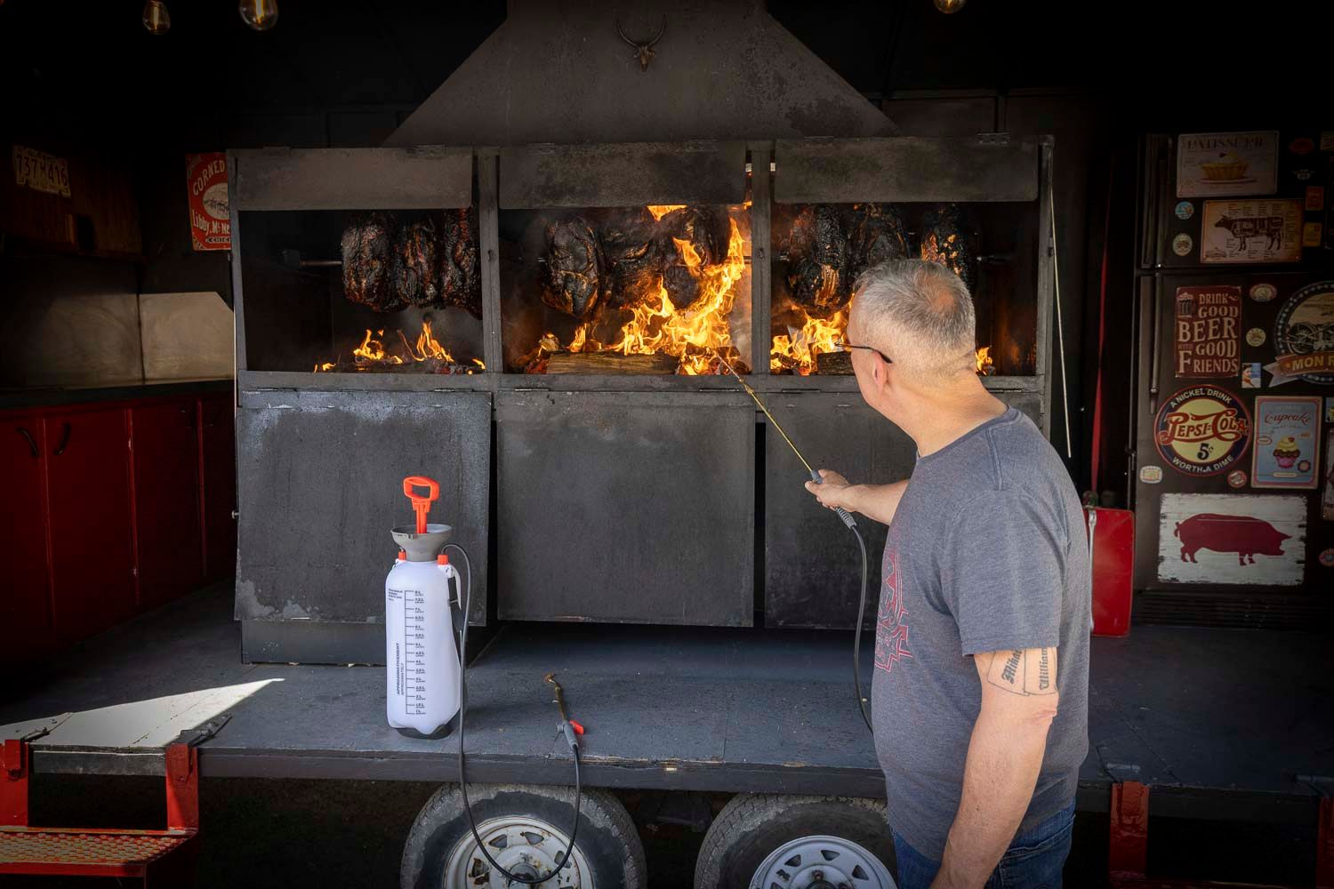 Man tending to open flame BBQ smoker trailer at outdoor cooking event.