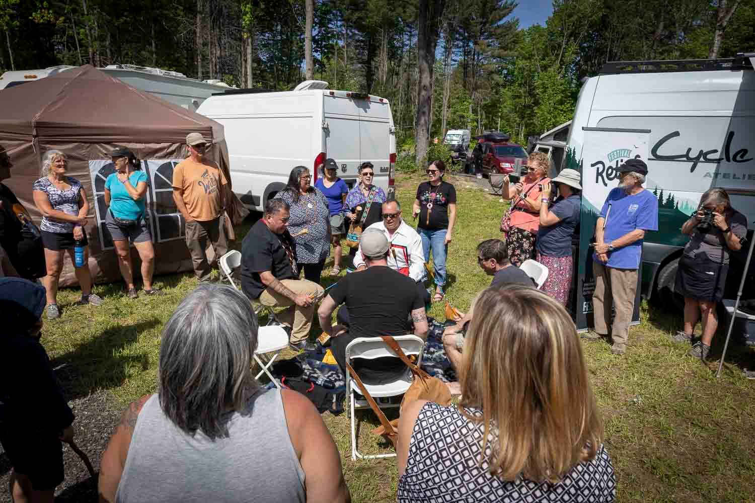 Group of people gathered outdoors near RVs and vans in wooded campground area on sunny day.