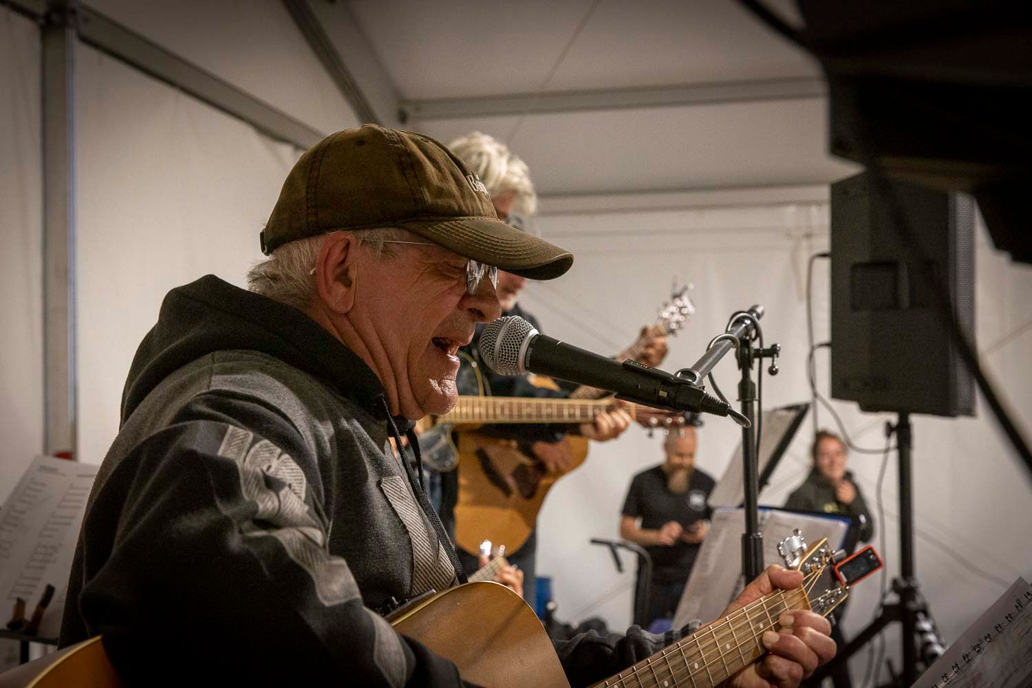 Older man in cap playing acoustic guitar and singing into microphone at outdoor music event.