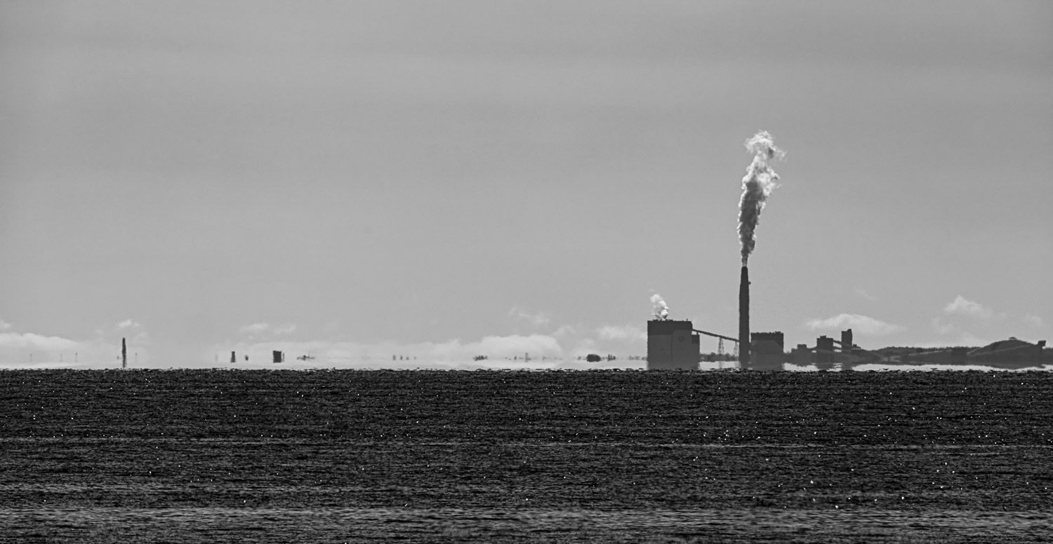 A black and white landscape of an industrial facility with a smoking chimney across a flat open field.