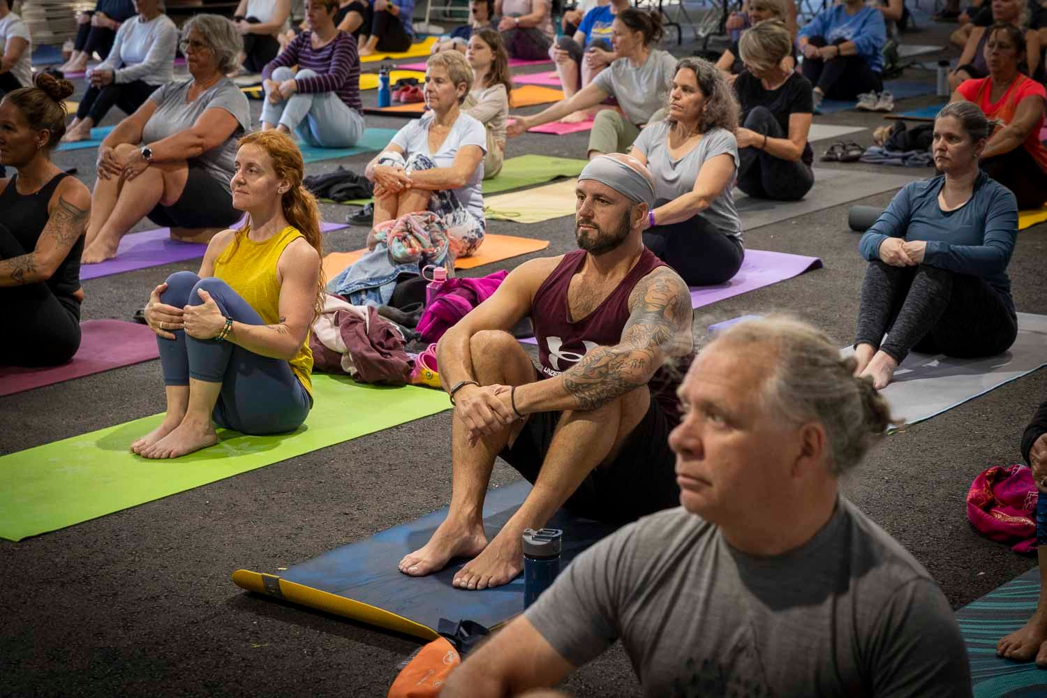 Large group of diverse adults sitting on colorful yoga mats during an indoor yoga class session.