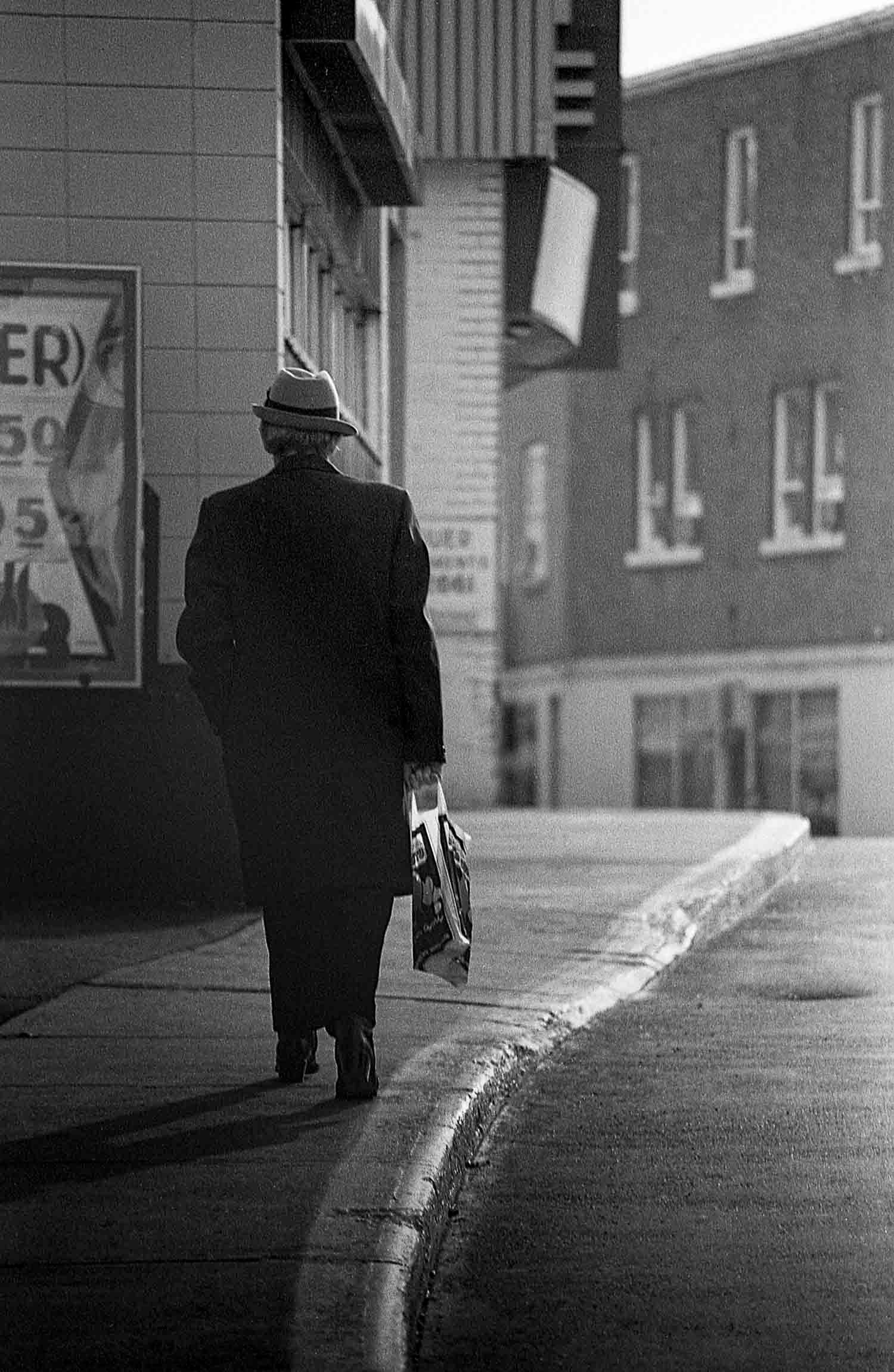 Homme de dos, qui marche
Black and white photo of an elderly person in a coat and hat walking alone on a sidewalk carrying a bag.