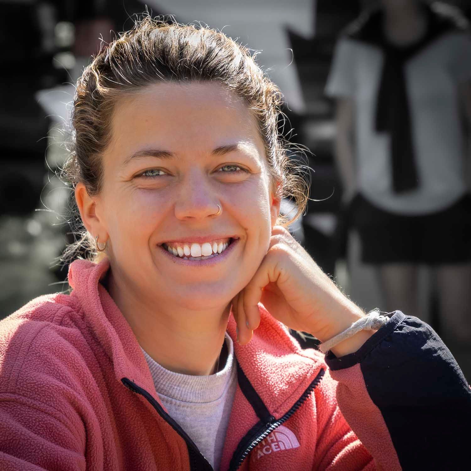 Smiling young woman with dark hair wearing a red North Face fleece jacket, sitting outdoors in bright sunlight.