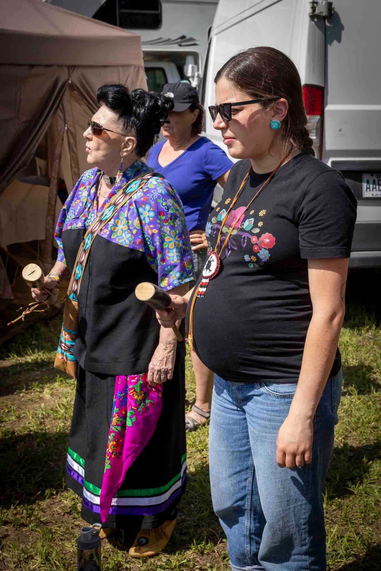 Two Indigenous women in traditional regalia holding ceremonial items at an outdoor cultural gathering.