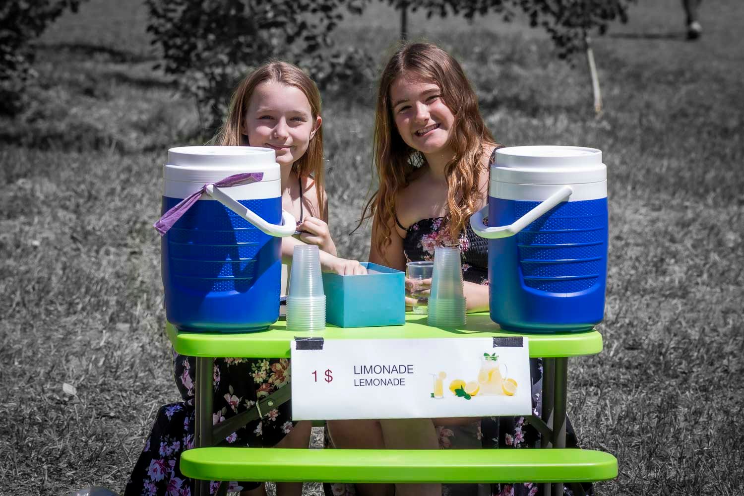 Two girls smiling behind lemonade stand with blue coolers selling drinks for $1.