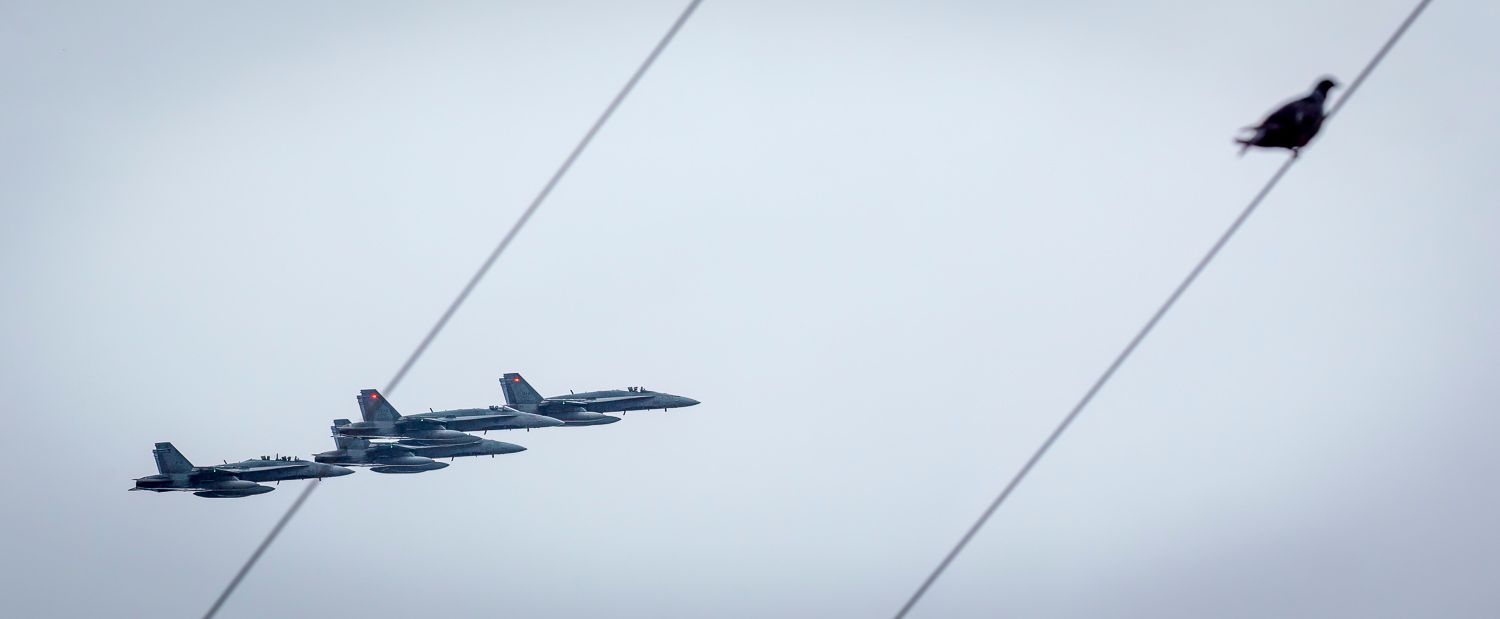 Three military fighter jets flying low in formation past a red flag against a grey sky.