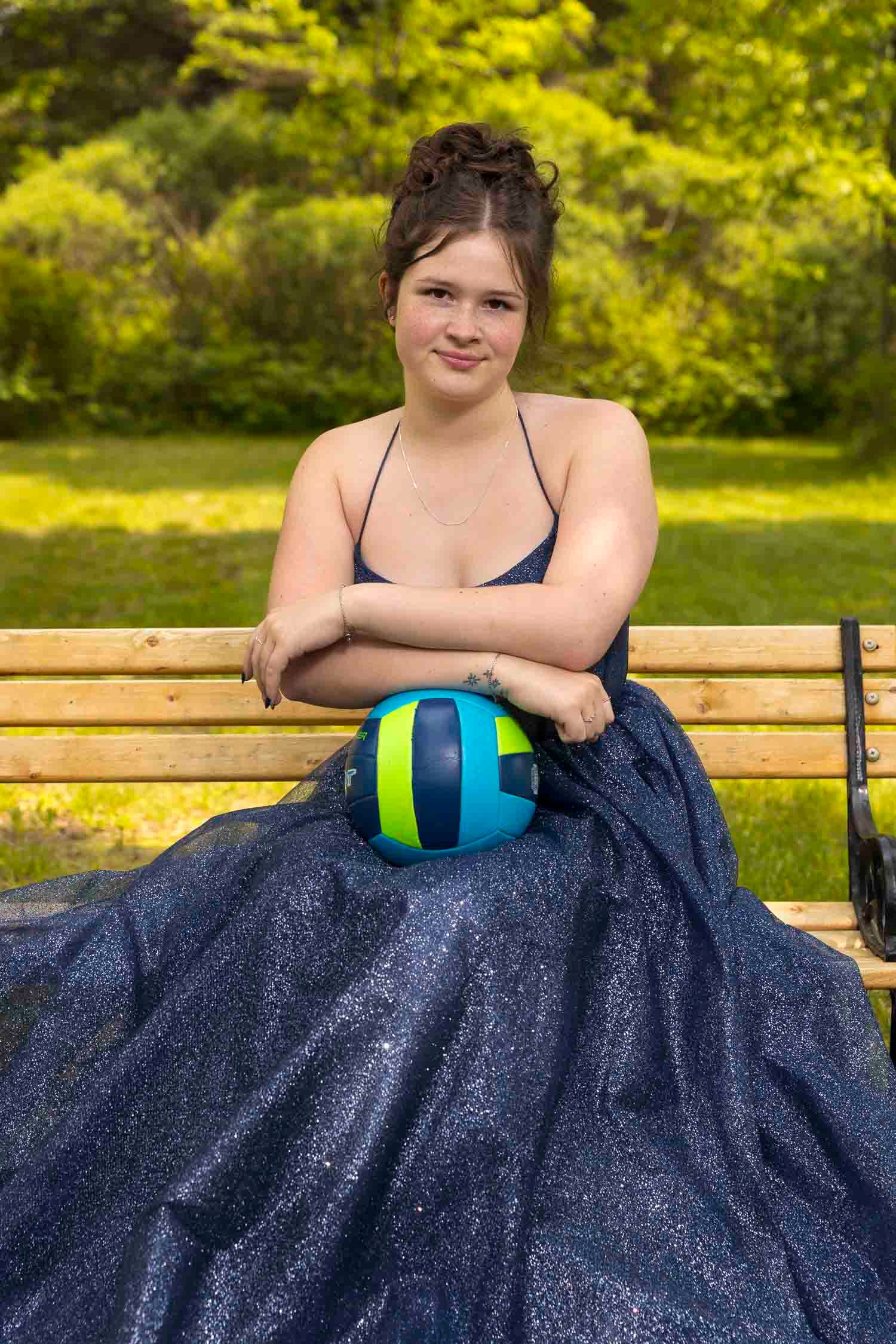 Young woman in a sparkling blue gown sitting on a park bench holding a volleyball outdoors.