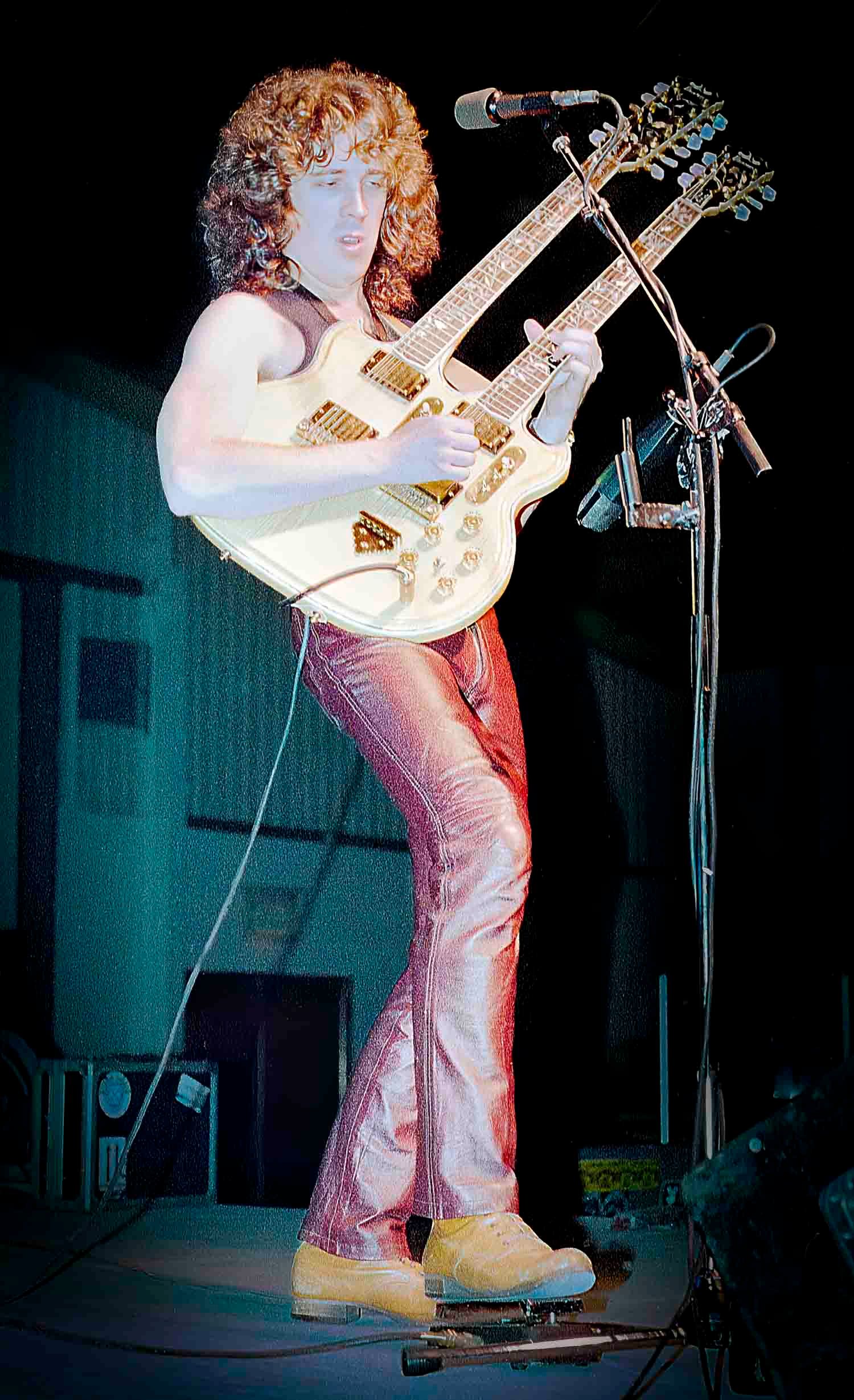 Curly-haired rock guitarist performing on stage with a white double-neck electric guitar wearing shirtless and red pants.