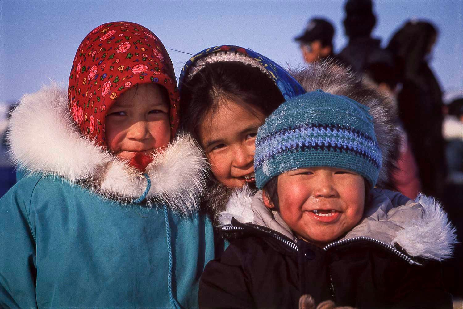 Enfants Inuits, Nunavik,
Three smiling Inuit children wearing colorful winter hats and fur-trimmed parkas outdoors in a cold Arctic setting.