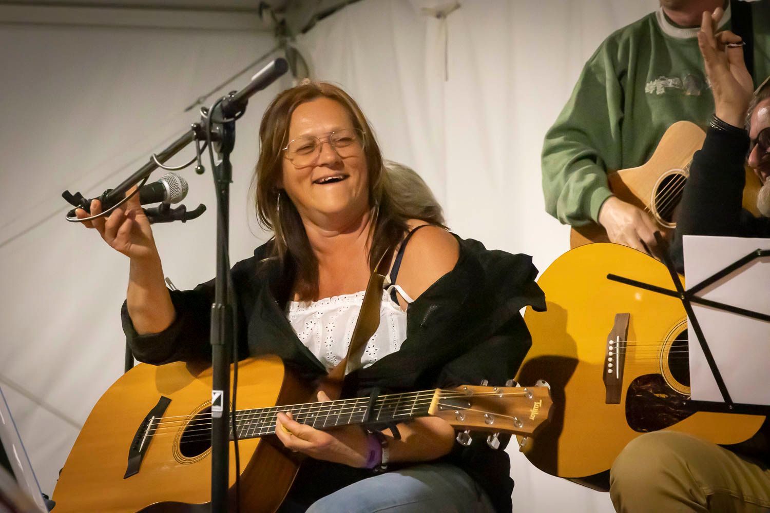 Smiling woman playing acoustic guitar on stage at an outdoor music event under a white tent.