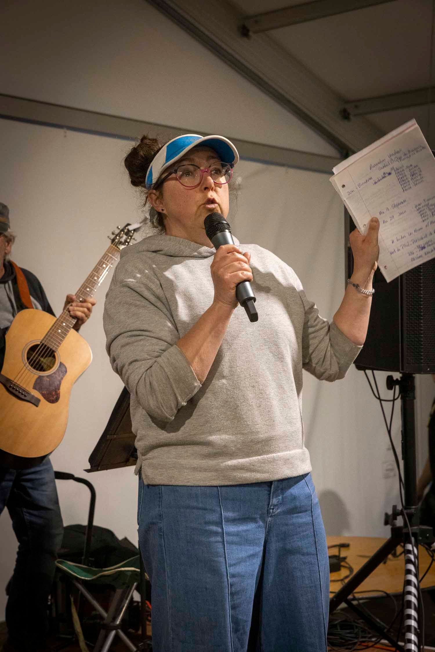 Woman in gray hoodie and blue cap speaks into microphone while holding papers at an outdoor music event.