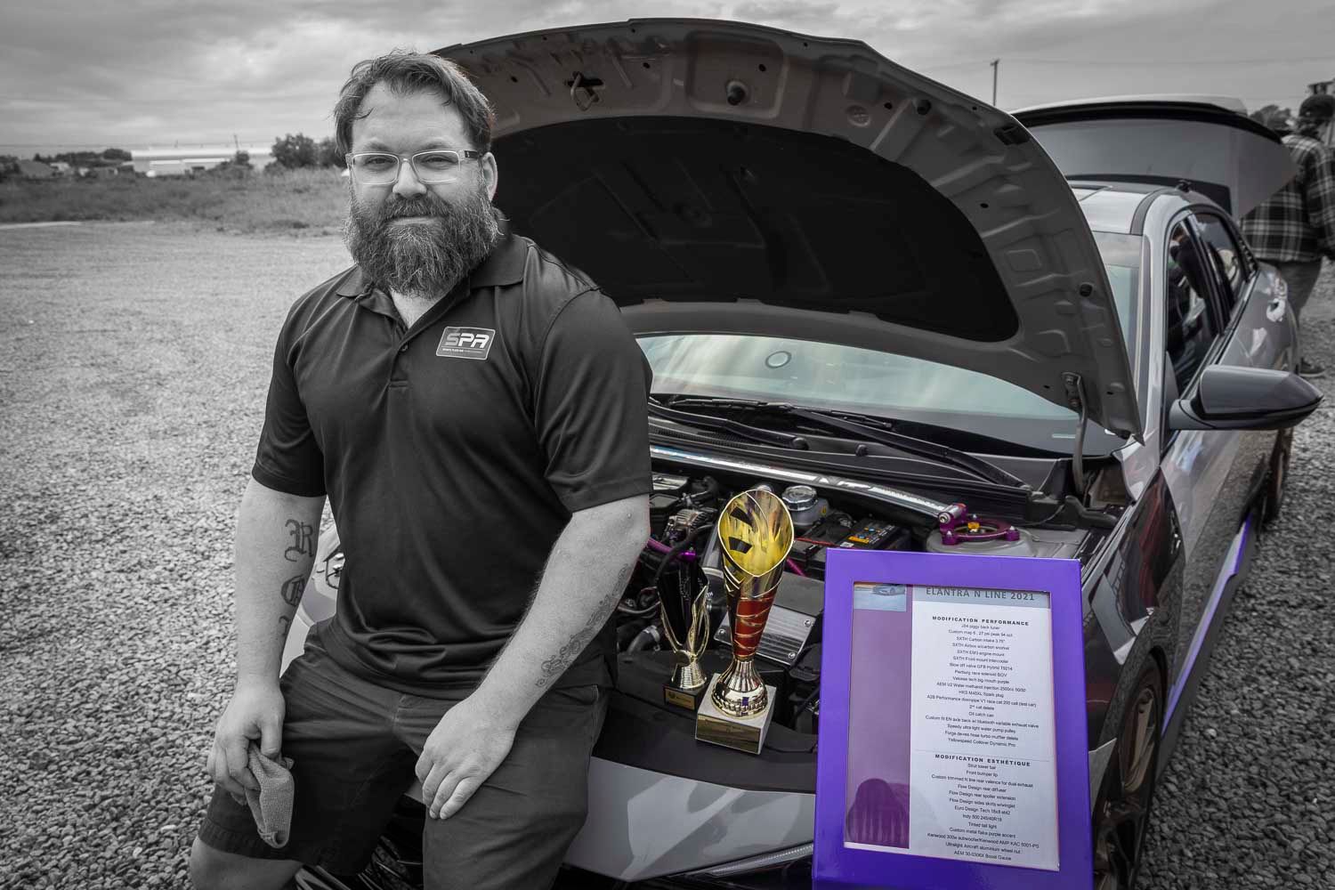 Bearded man posing proudly beside a car with open hood displaying engine and trophy at an outdoor automotive show event.