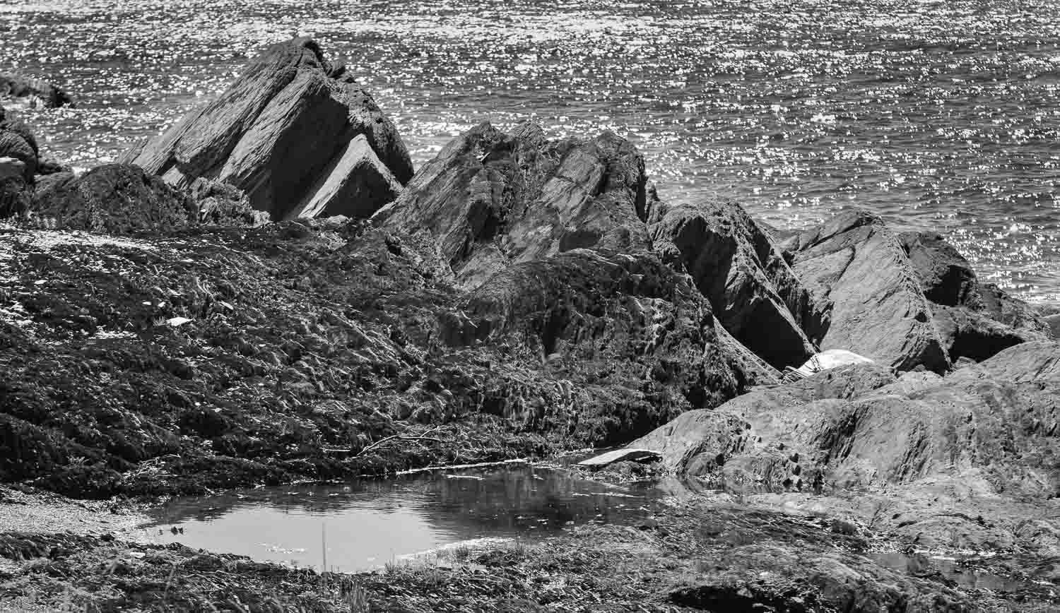 Black and white photo of rocky coastal landscape with small tidal pool among weathered boulders and rugged terrain.