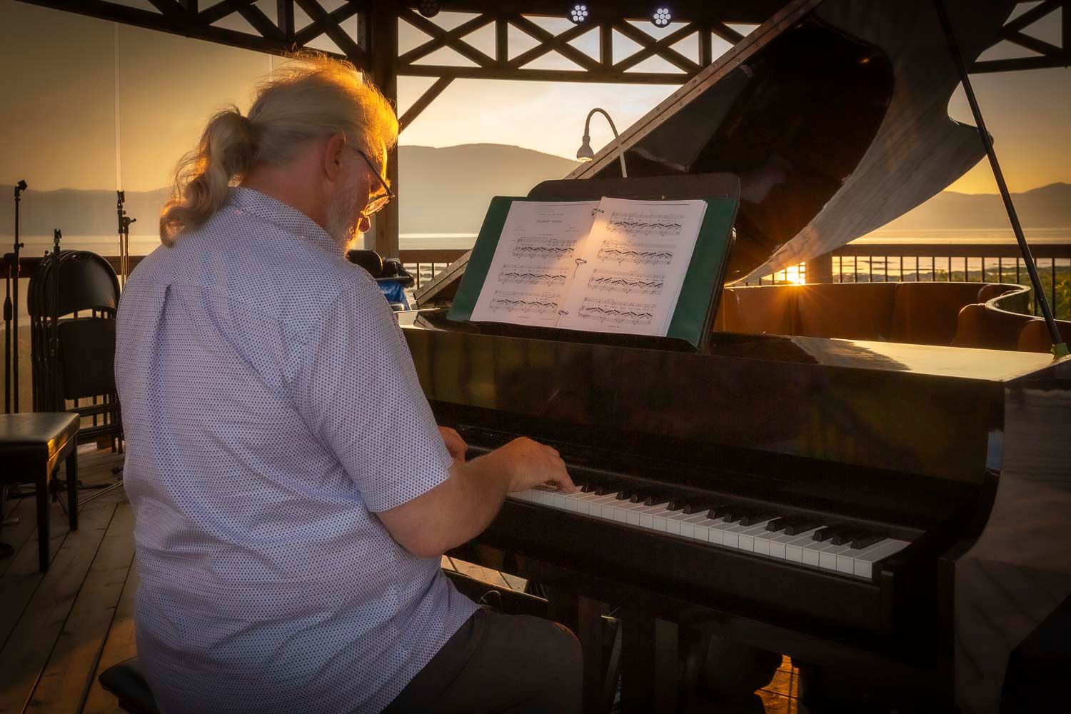 A musician with long hair plays a grand piano outdoors at sunset with sheet music on the stand.