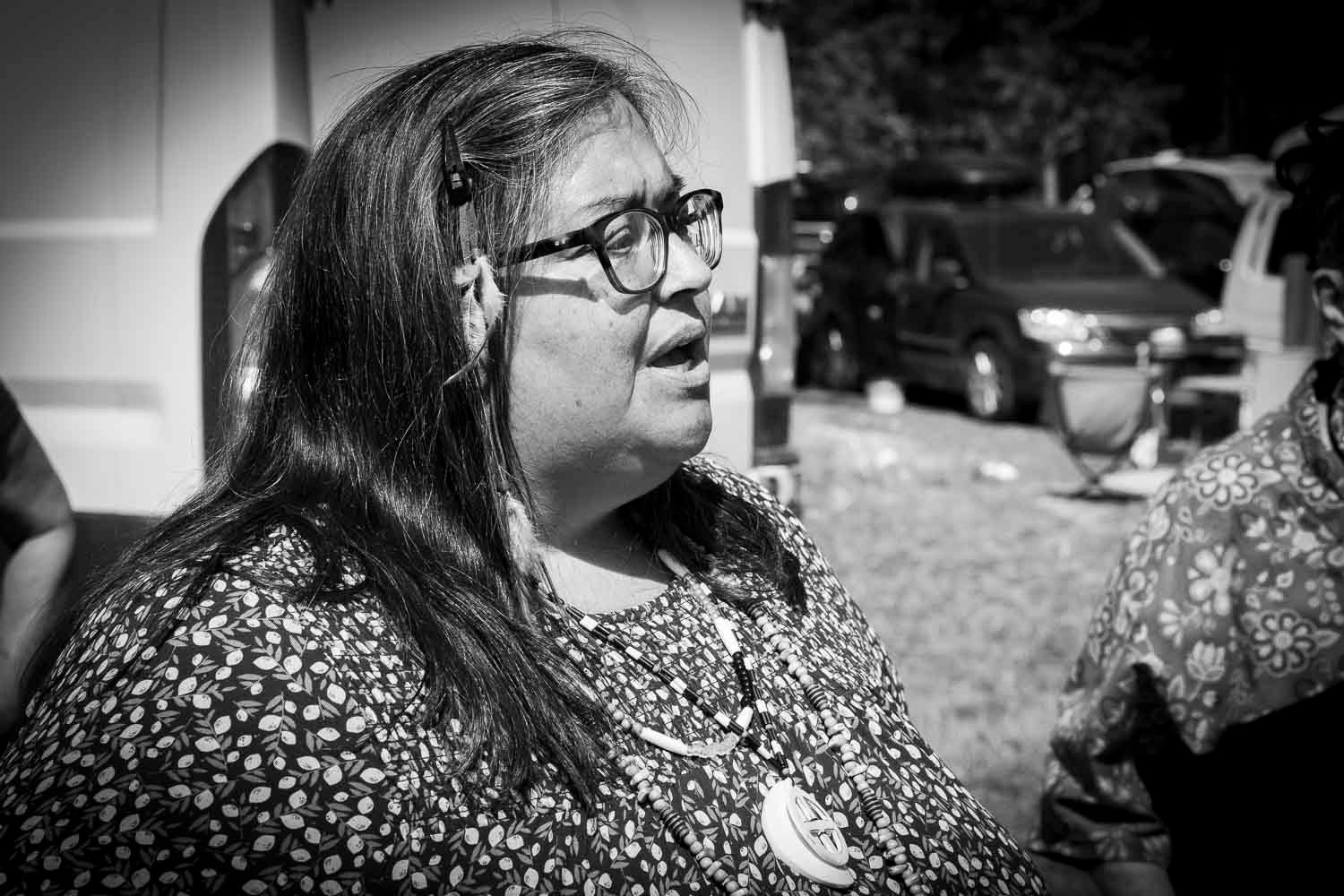 Black and white portrait of a woman with glasses and long hair at an outdoor event.