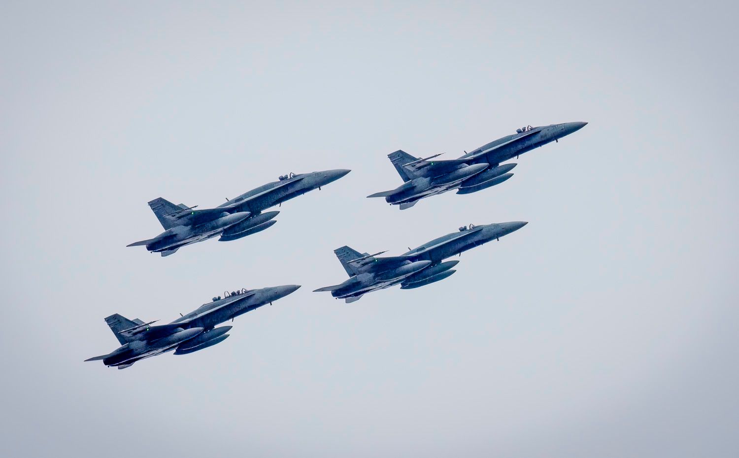 Four blue and yellow CF-18 Hornet fighter jets flying in tight diamond formation against grey sky.