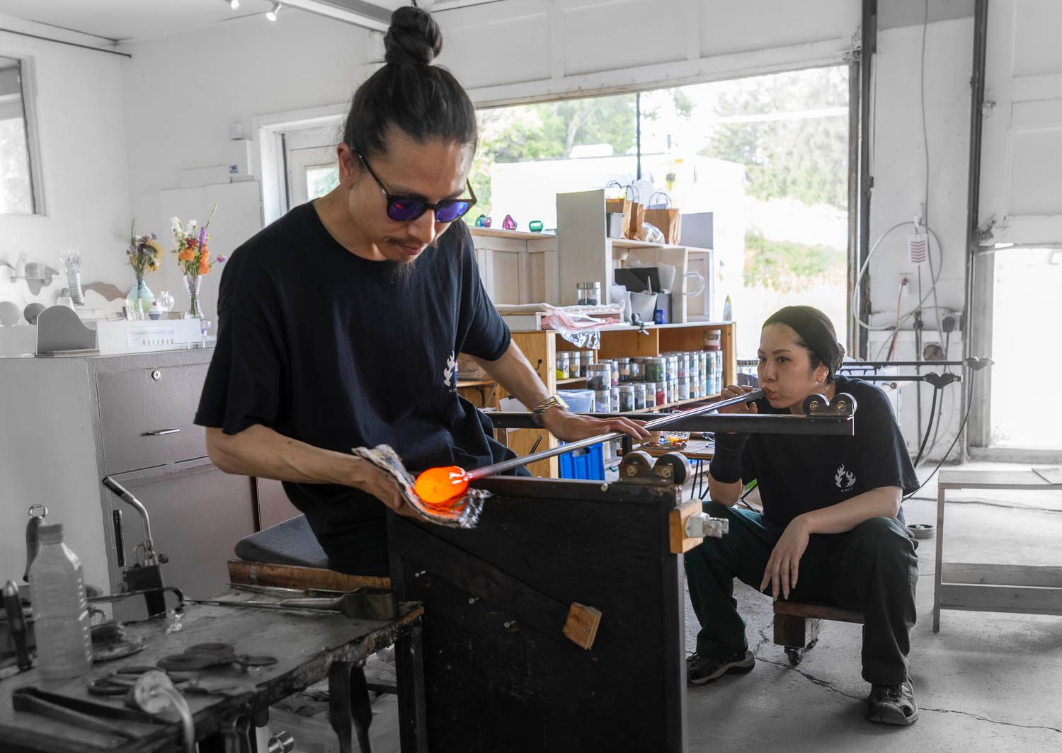 Glassblower shaping molten glass in a bright studio workshop while an observer watches nearby.
