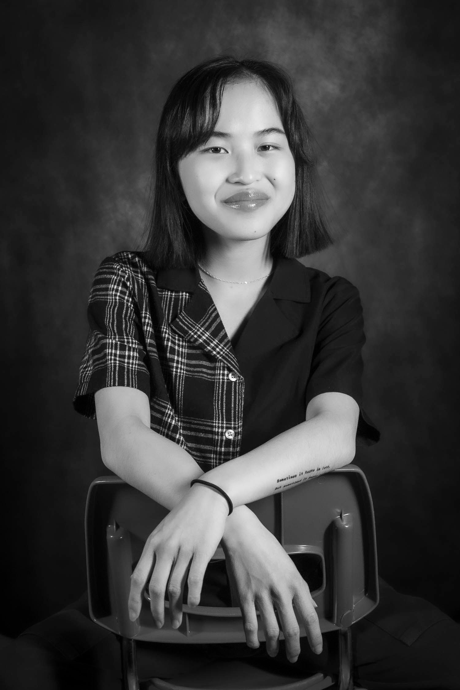 Black and white studio portrait of a smiling young Asian woman sitting relaxed on a chair with arms crossed.