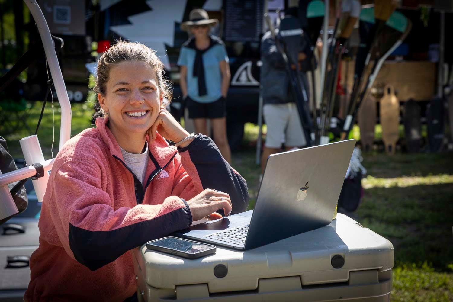 Smiling woman in pink jacket working on laptop outdoors at a sunny event with people and equipment in background.