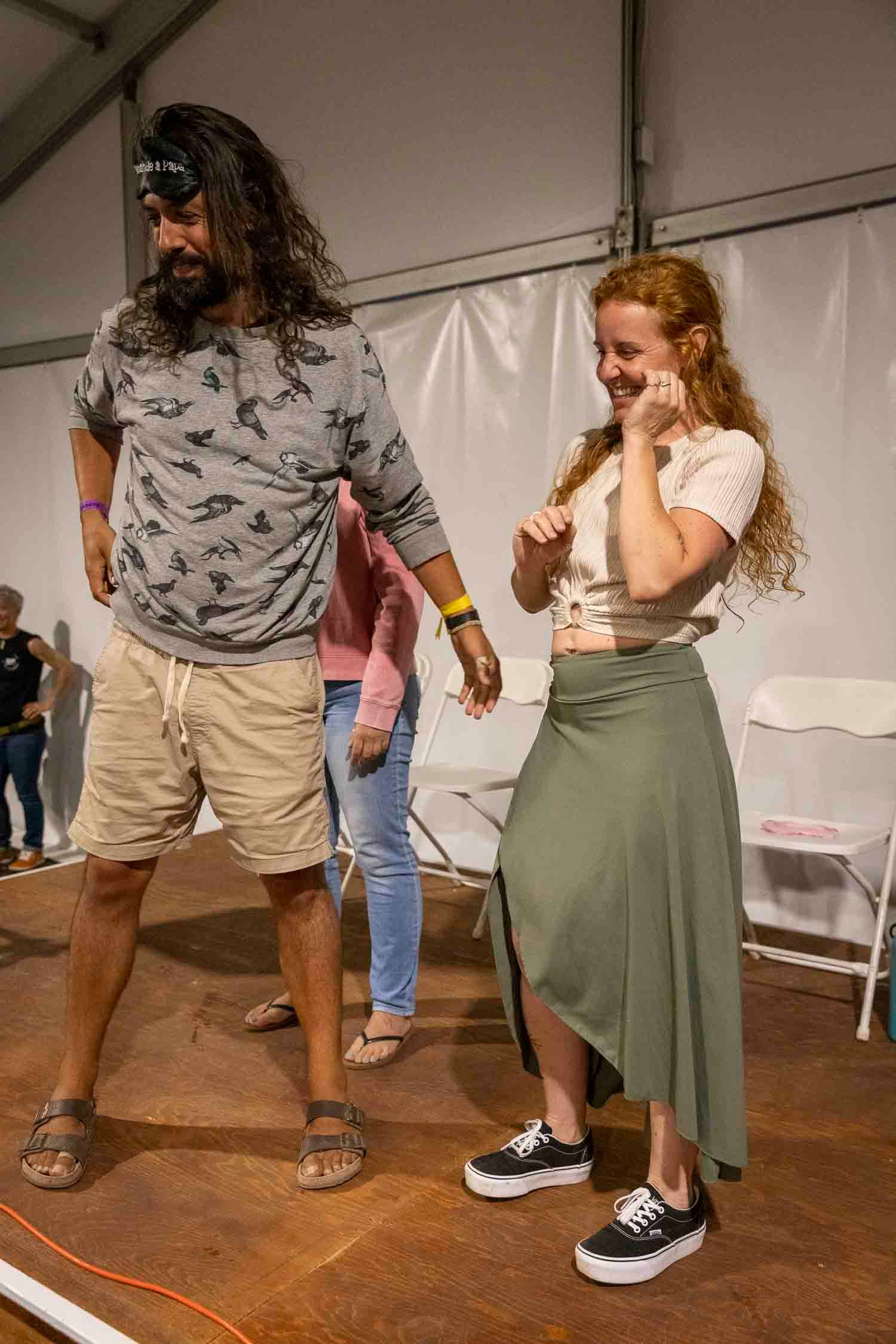 Man and woman dancing and laughing together on a stage at an outdoor event under a tent.