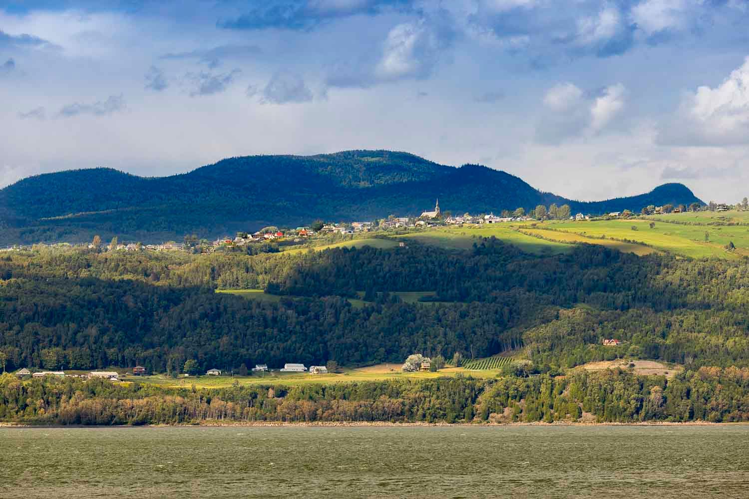 Les Éboulements, QC, Canada
A picturesque village sits atop rolling green hills covered with dense forests under a cloudy sky.
