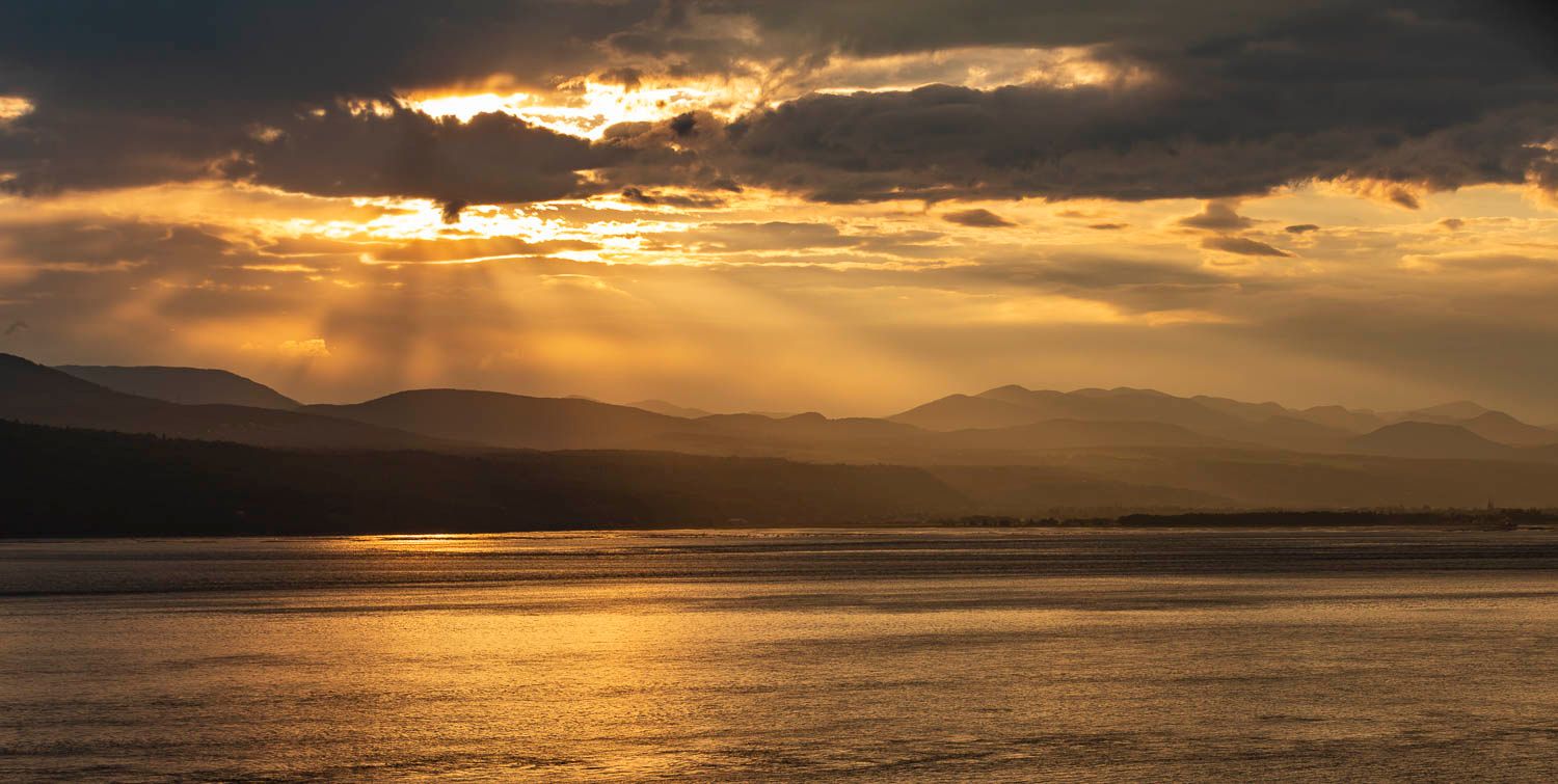 Golden sunset over calm water with dramatic rays piercing through clouds above silhouetted mountains.