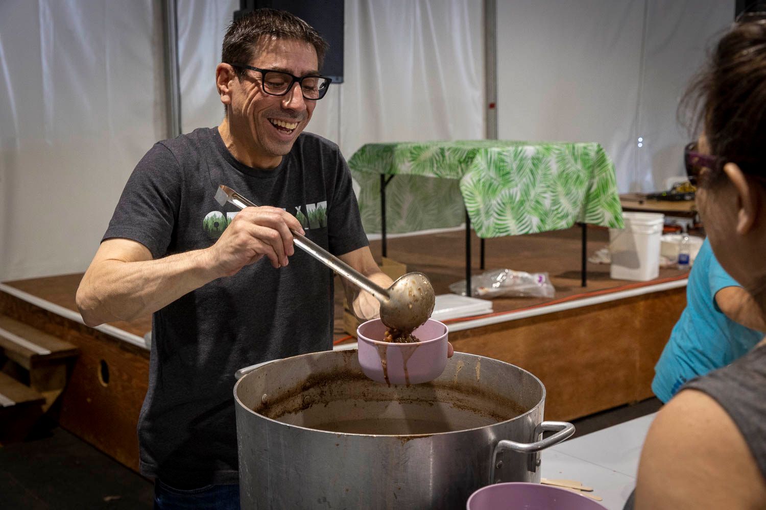 Smiling man ladling soup from a large pot into a bowl at an outdoor food event.