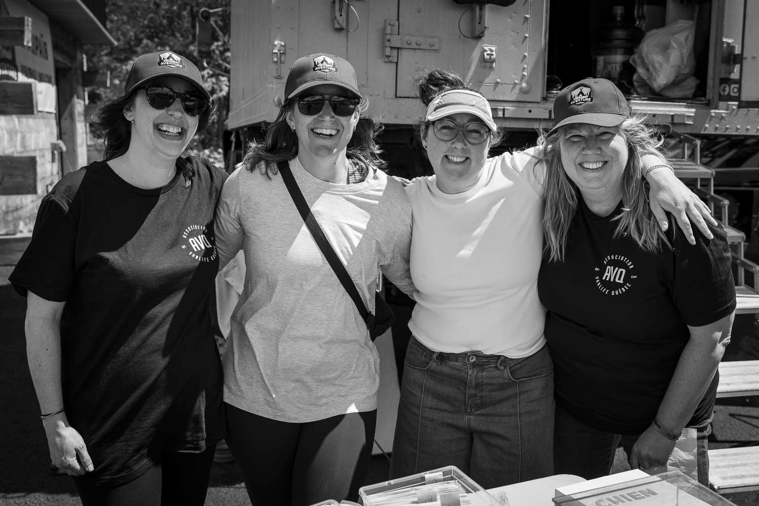 Four smiling women pose together outdoors at a community event wearing matching black shirts and caps.