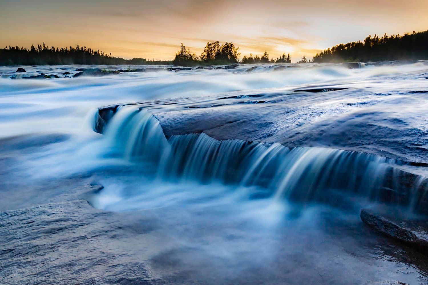 Chute à l'ours, Normandin.
Long exposure waterfall cascading over rocky ledges at sunset with misty blue water and golden sky.