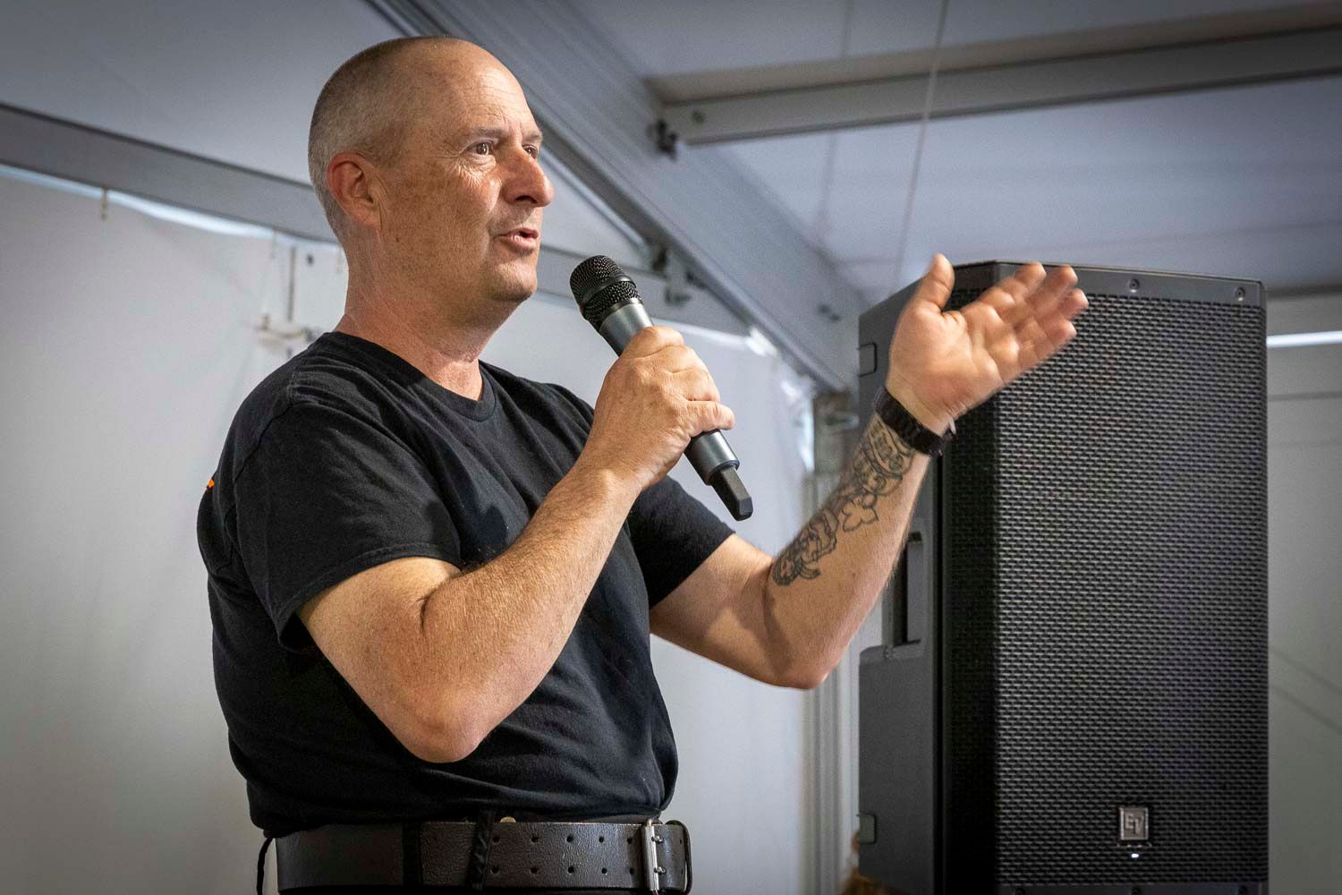 Tattooed man in black shirt speaking into a microphone at an indoor event with audio speakers nearby.